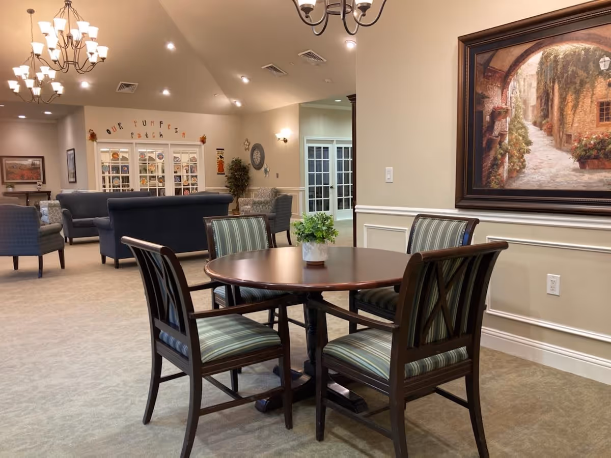 A cozy common area in a senior living facility featuring a round wooden table with four striped cushioned chairs. In the background, there are several upholstered sofas and armchairs arranged for seating. The walls are decorated with framed artwork and a sign that reads 'Our Pumpkin Patch'. The room is softly lit with chandeliers and wall sconces, and there is a potted plant on the table and another near the wall.
