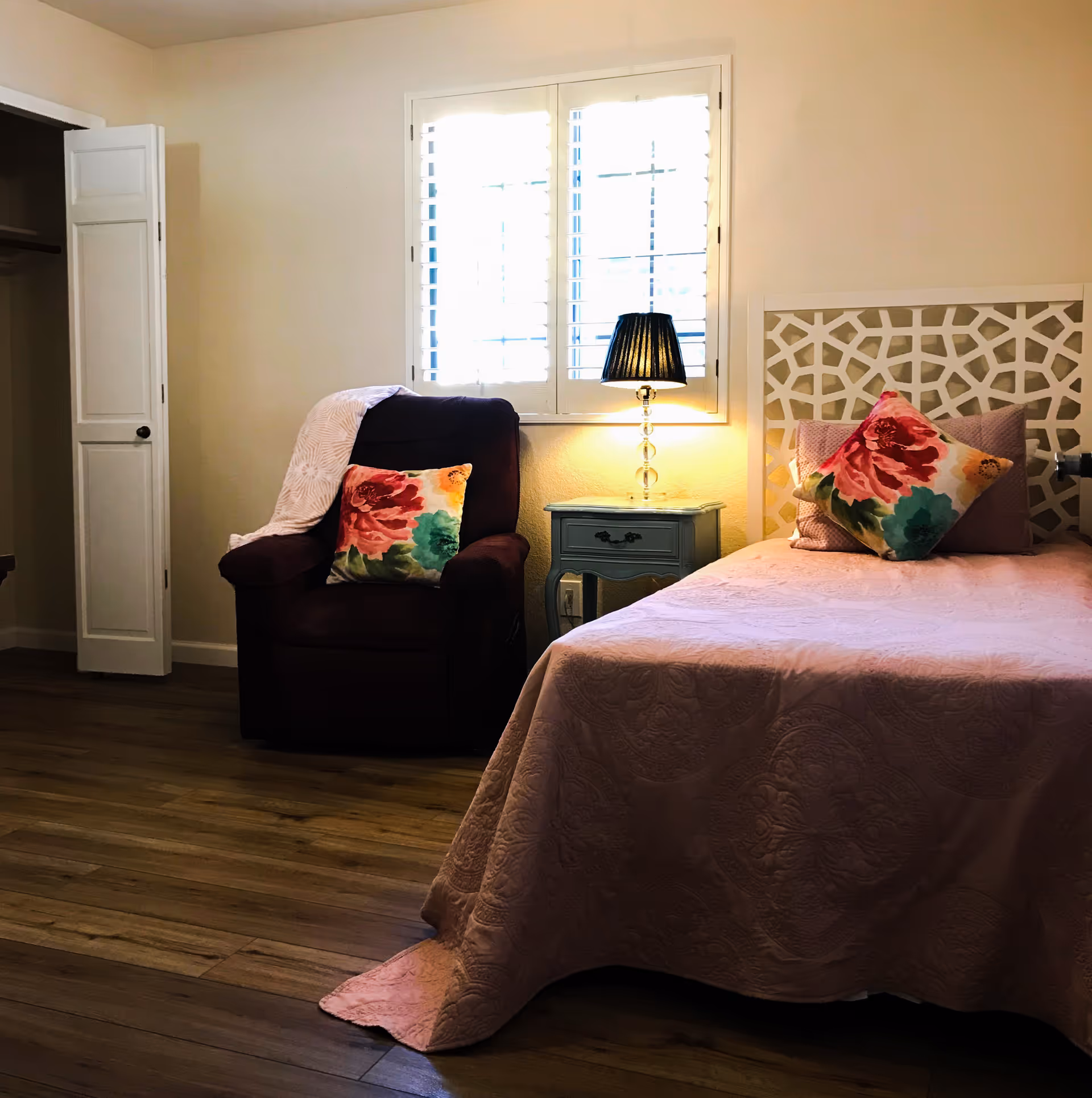 A cozy bedroom with a single bed covered in a pink quilt and decorated with floral pillows. Next to the bed is a small blue nightstand with a lamp that has a black shade. To the left of the nightstand is a dark armchair with a floral pillow and a light-colored blanket draped over the back. The room has wooden flooring and a window with white shutters behind the nightstand and armchair. An open closet door is visible on the far left side of the room.