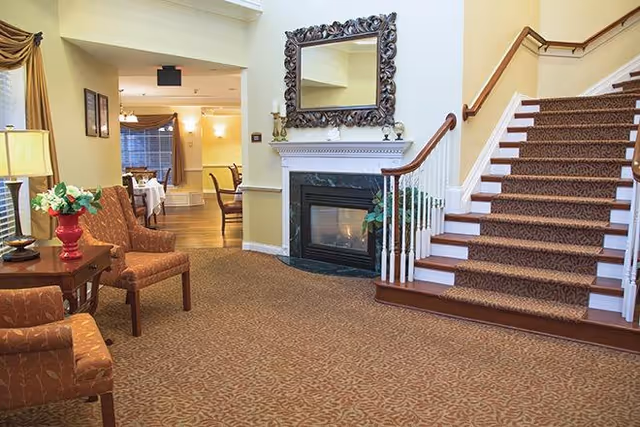 A cozy interior space featuring a carpeted staircase with a patterned runner, a fireplace with a decorative mirror above it, two upholstered chairs with a small table holding a lamp and a vase of flowers, and a dining area visible in the background.