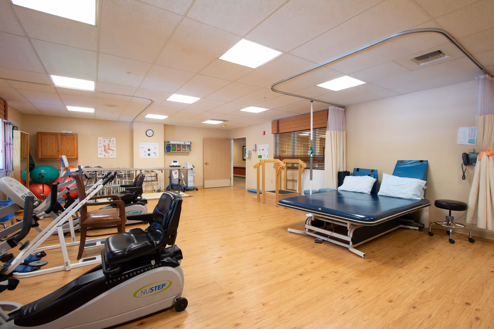 Rehabilitation therapy room with exercise machines, treatment table, parallel bars and therapy equipment on a wood floor.
