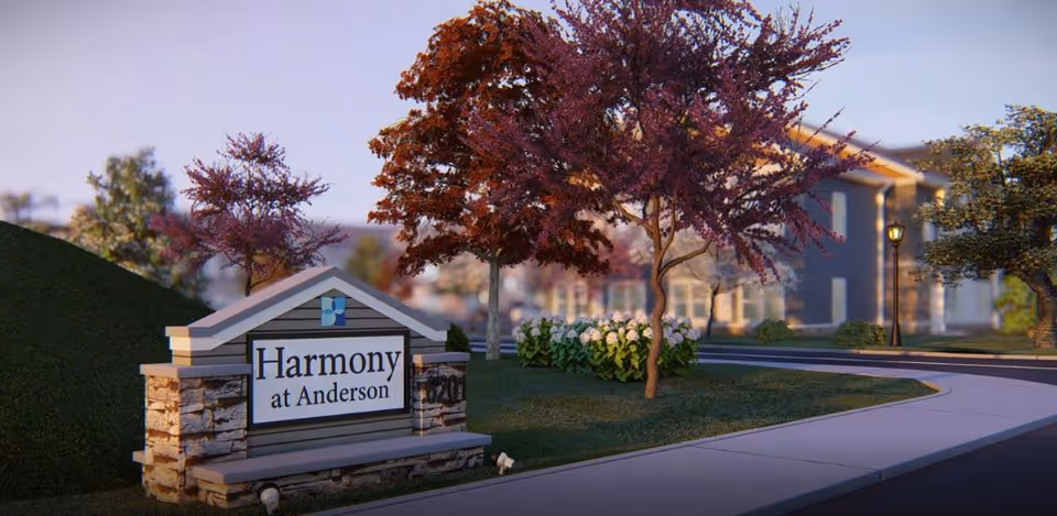 Entrance sign for Harmony at Anderson senior living facility surrounded by landscaping with trees and flowers, with a building and streetlamp visible in the background.
