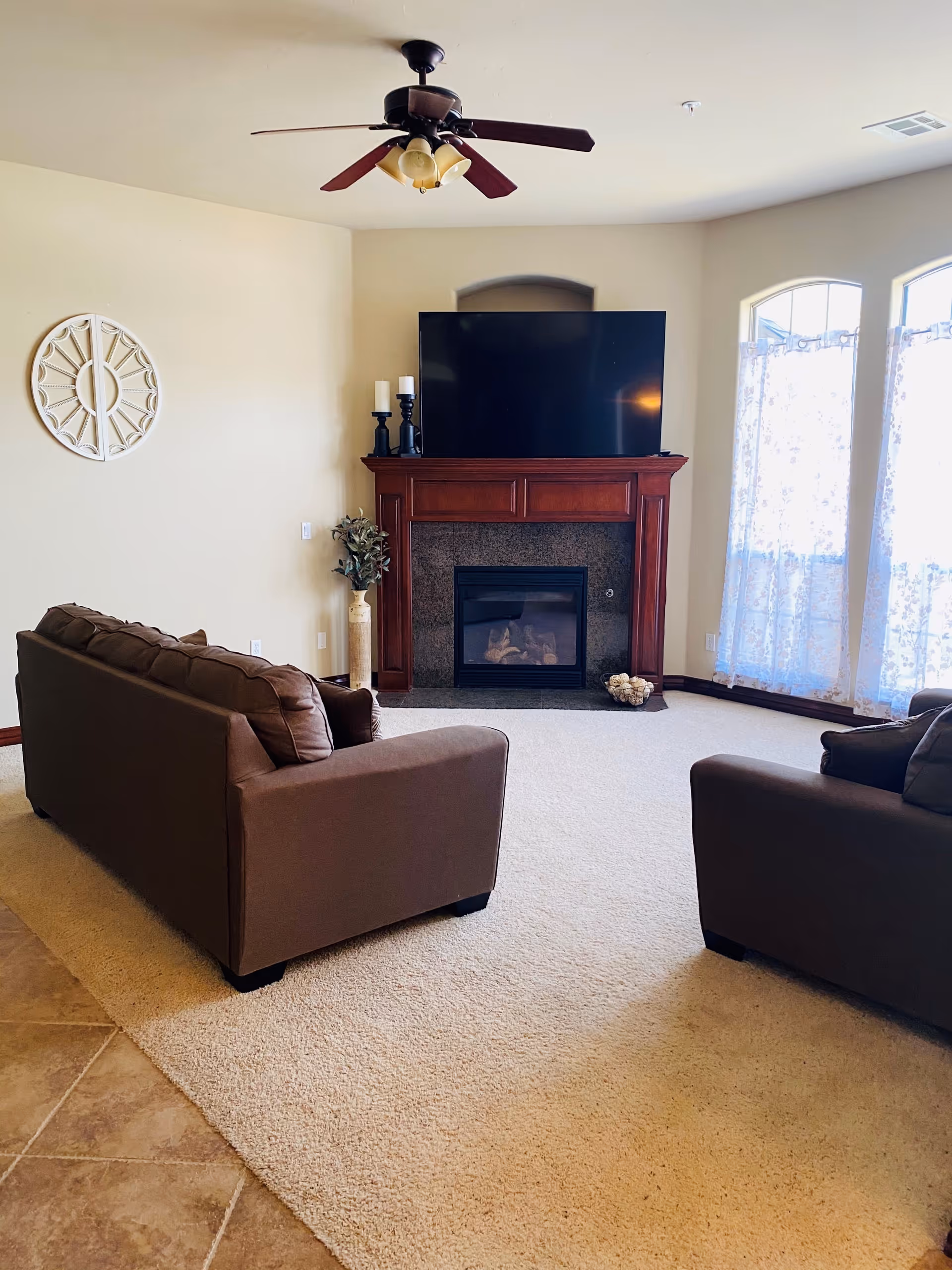 A living room with beige carpet and tiled floor, featuring two brown sofas facing a wooden fireplace with a large flat-screen TV mounted above it. There are three tall windows with sheer floral curtains on the right side, a ceiling fan with lights above, and decorative items including candles and a vase with greenery near the fireplace.