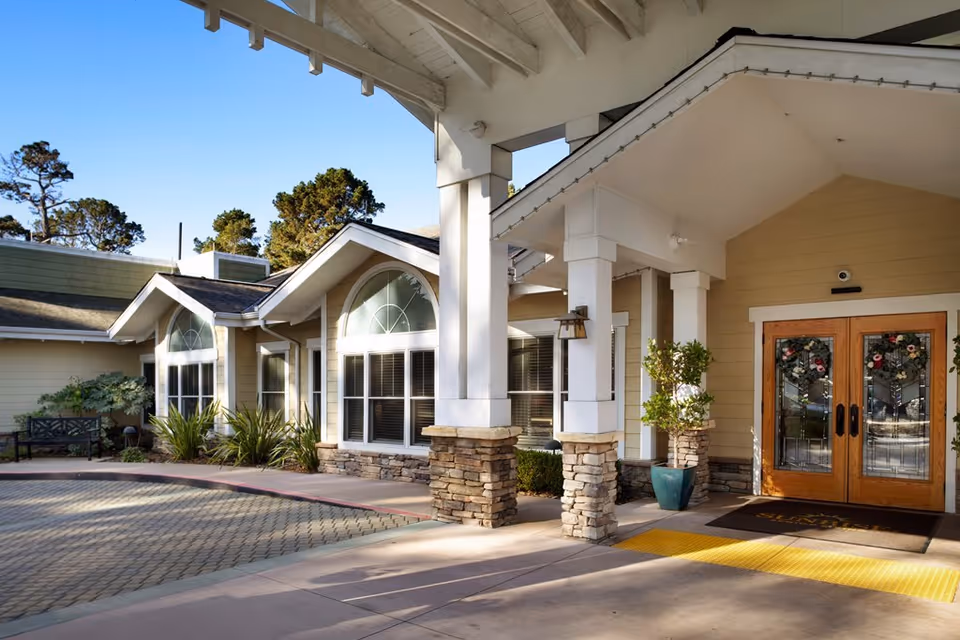 Exterior view of the entrance to a senior living facility with beige siding, stone pillars, large windows with white trim, and double wooden doors decorated with floral wreaths. There is a covered walkway with a yellow tactile paving strip and some greenery around the entrance.