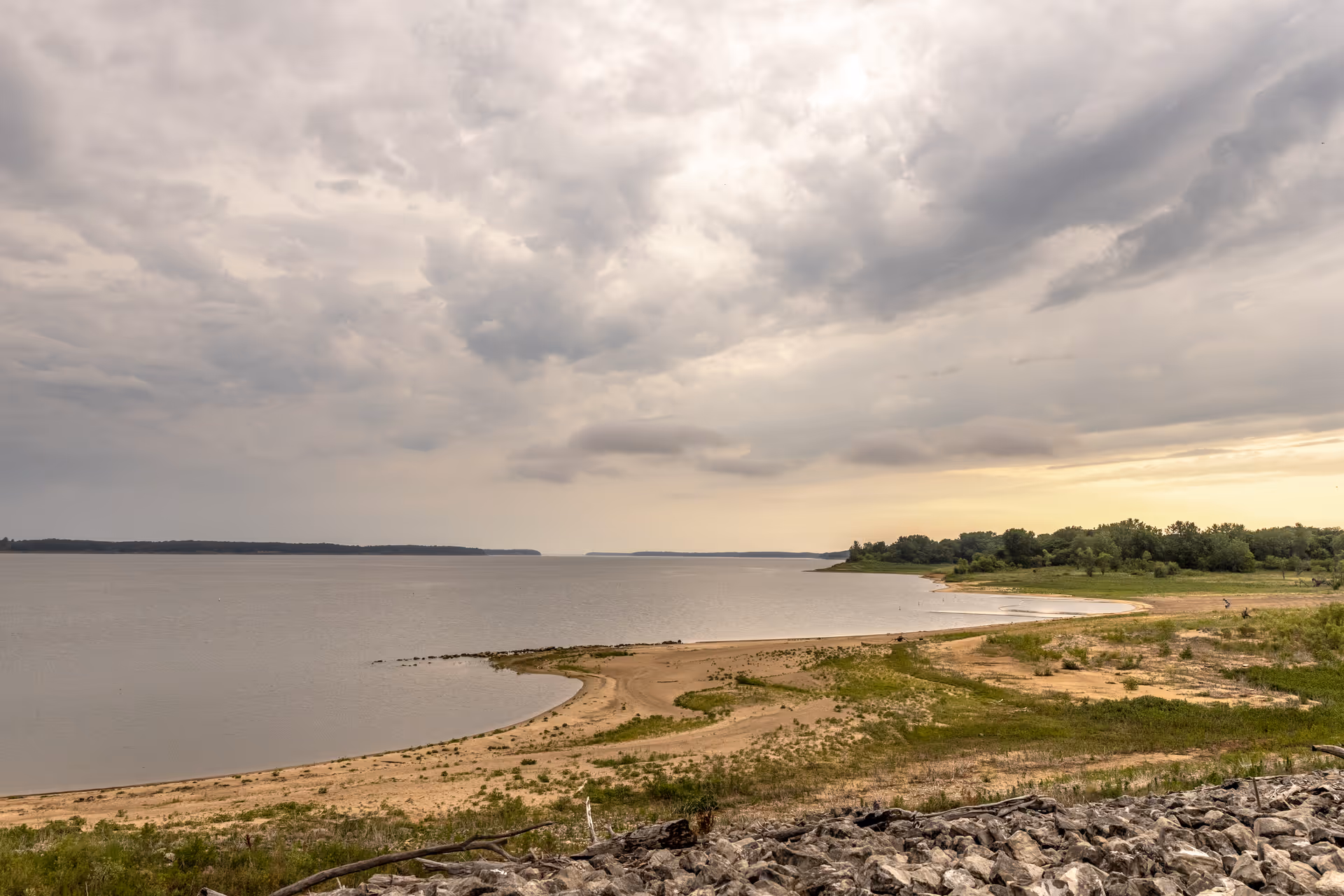 A wide view of a lakeshore with a sandy beach and patches of green vegetation under a cloudy sky during sunset or sunrise.