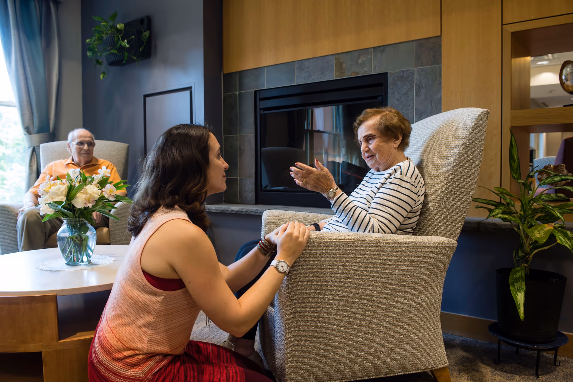 A young woman kneeling and holding hands with an elderly woman seated in a beige armchair in a cozy living room with a fireplace. An elderly man sits in another armchair near a window with curtains, and a table with a vase of flowers is between them. There is a potted plant on the right side of the image.