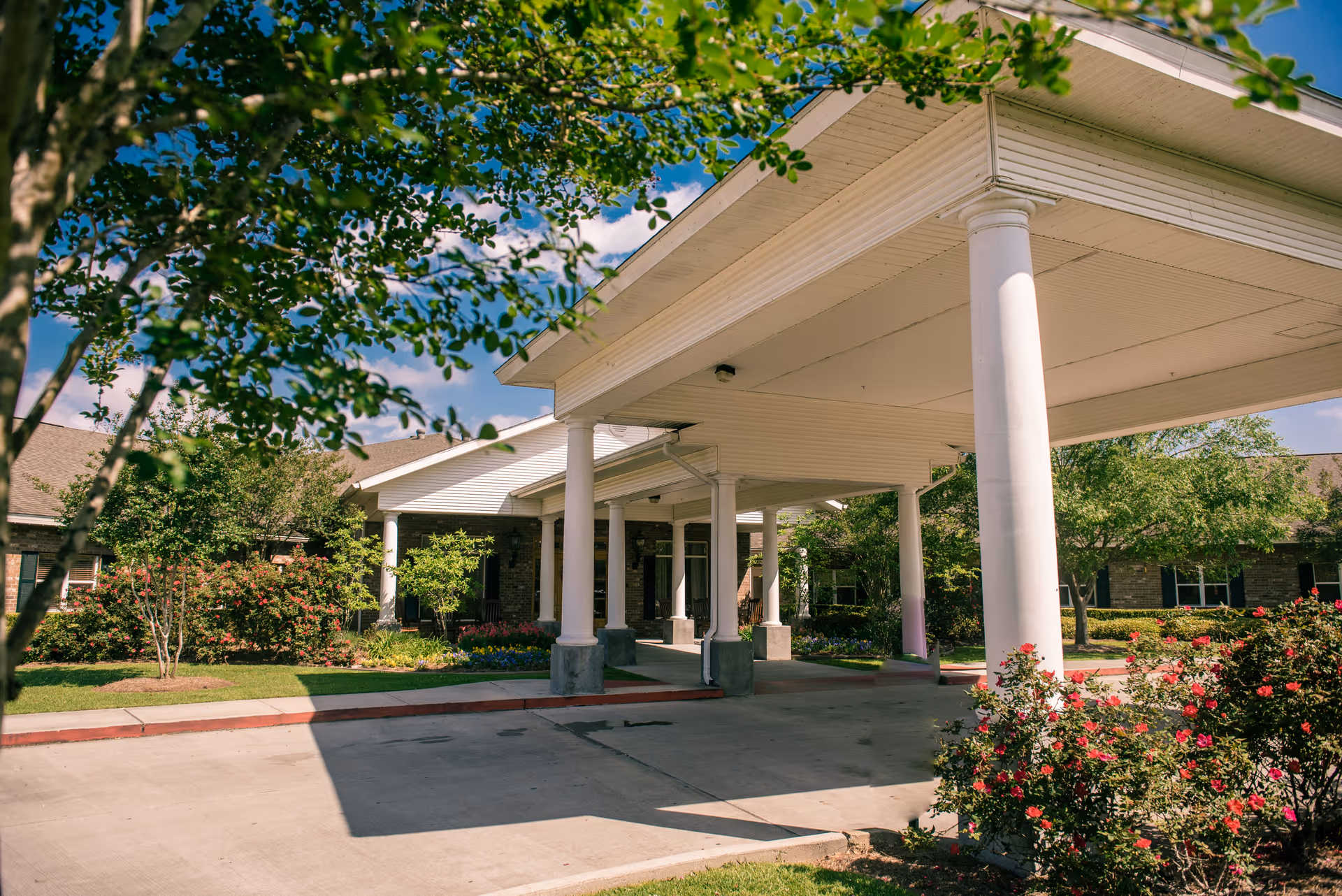 Covered porte-cochère with white columns and landscaped gardens at the front entrance of the facility.