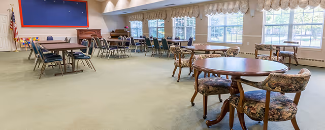 Bright, carpeted community dining/activity room with multiple round and rectangular tables and chairs and large windows along one wall.