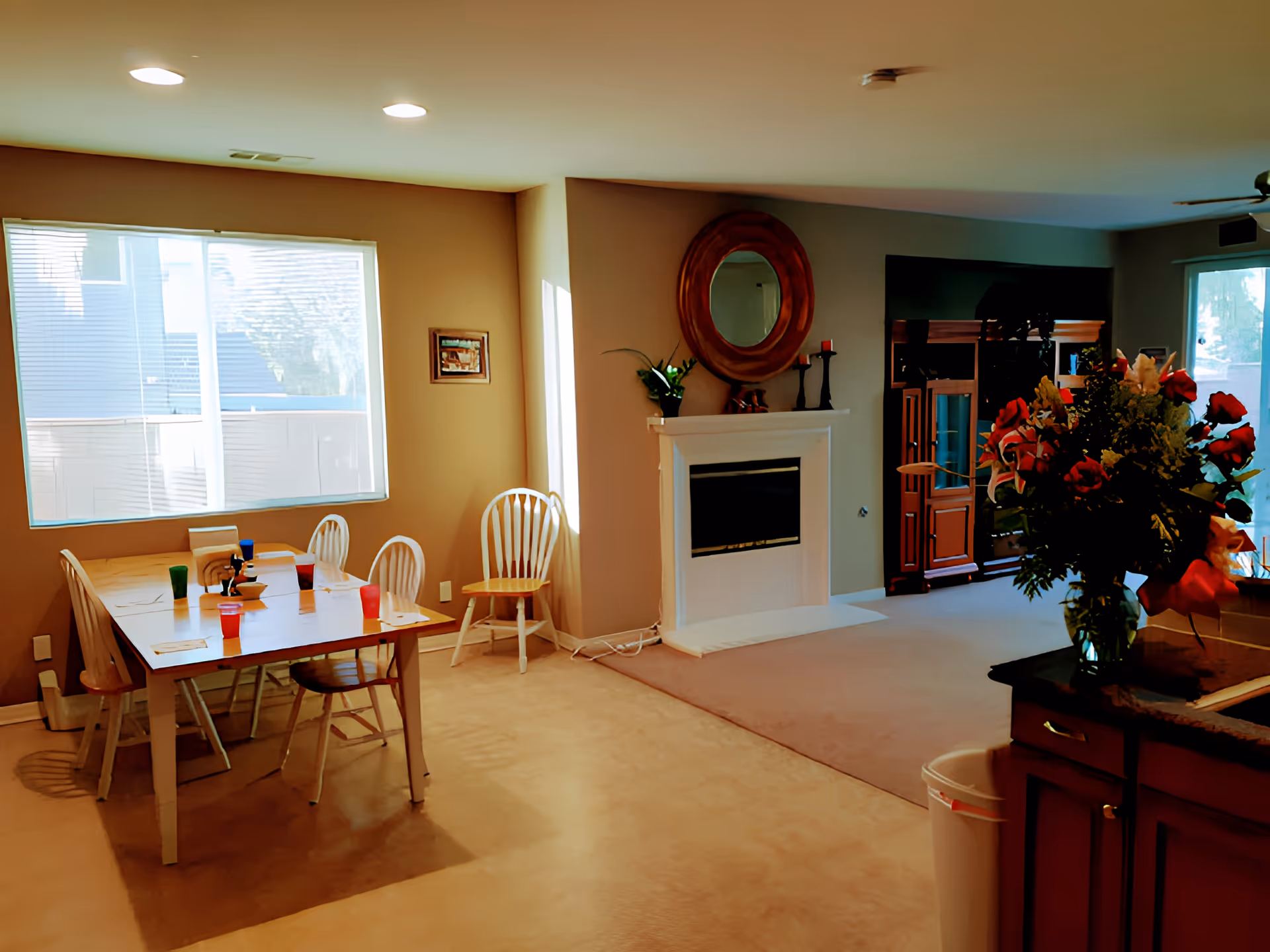 A cozy interior space featuring a dining area with a wooden table and six chairs, each with a colorful cup on it. Large windows allow natural light to brighten the room. Adjacent to the dining area is a living space with a white fireplace adorned with a round mirror and decorative items. A wooden entertainment center is visible in the background, and a vase with a bouquet of flowers sits on a countertop in the foreground.