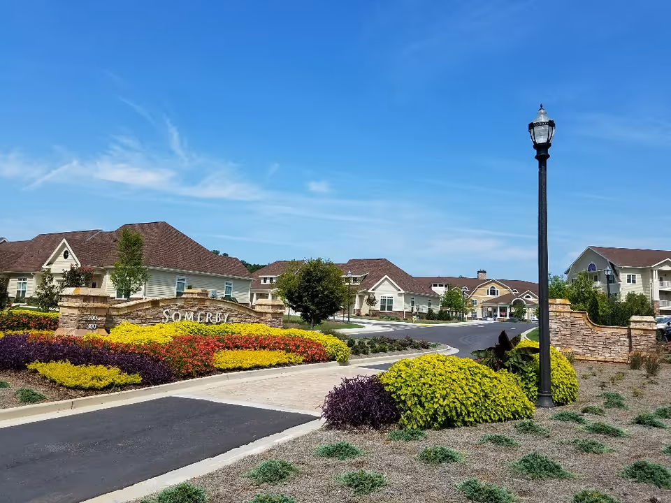 Entrance to Somerby Peachtree City senior living facility with a stone sign surrounded by colorful landscaping including yellow, red, and purple plants. Several beige buildings with brown roofs are visible in the background under a clear blue sky. A black street lamp stands near the entrance.
