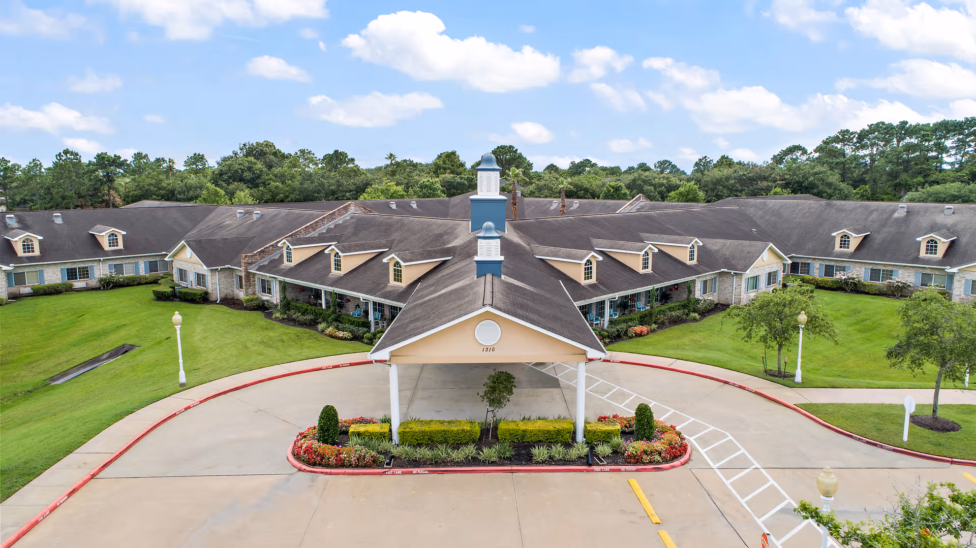 Front exterior view of Emeritus at Friendswood, a single-story senior living facility with a large covered entrance, surrounded by well-maintained green lawns, trees, and flower beds under a partly cloudy sky.