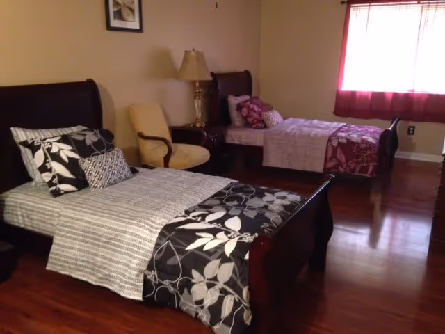 A bedroom with two single beds, each with patterned bedding and multiple pillows. There is a wooden nightstand between the beds with a lamp on it. A beige armchair is placed next to the nightstand. The room has wooden flooring and a window with red curtains letting in natural light.
