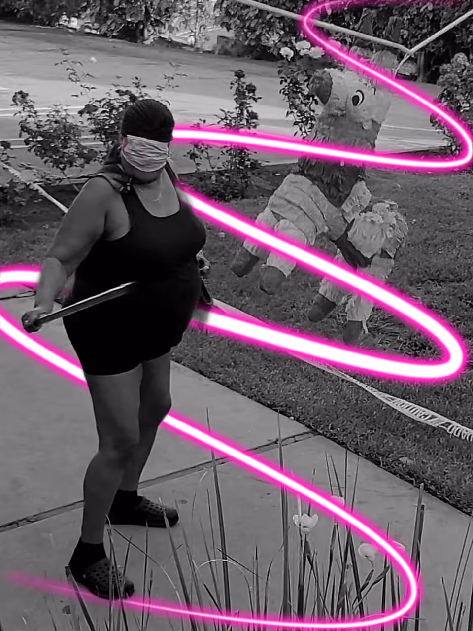 A person wearing a blindfold and holding a stick is standing on a sidewalk outdoors, preparing to hit a piñata shaped like a donkey. The image is black and white with bright pink neon light trails spiraling around the person and the piñata. There are plants and grass visible in the background.