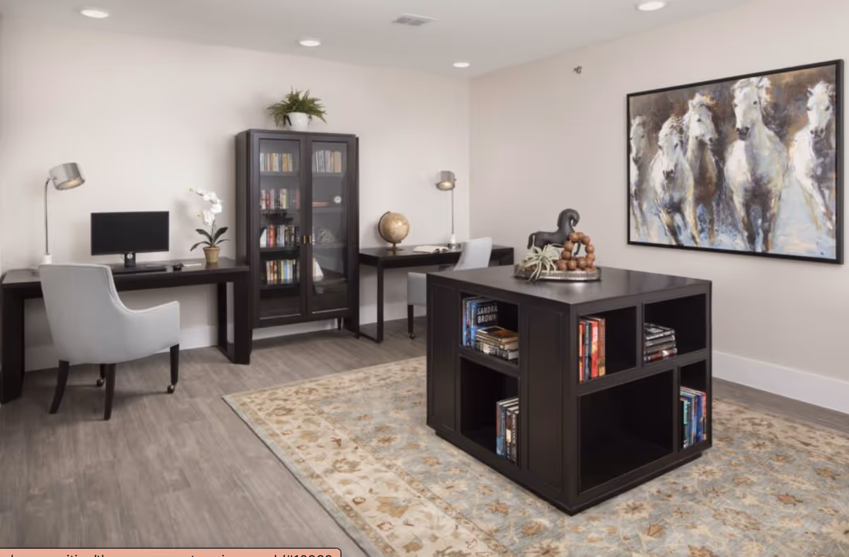 A well-lit room with two desks, each with a chair and desk lamp. One desk has a computer monitor and a potted orchid, while the other has a globe and an open book. Between the desks is a glass-front bookcase filled with books. In the center of the room is a dark wooden cube-shaped bookshelf with books and decorative items on top. A large painting of white horses hangs on the wall. The floor has a light-colored patterned rug over wood flooring.