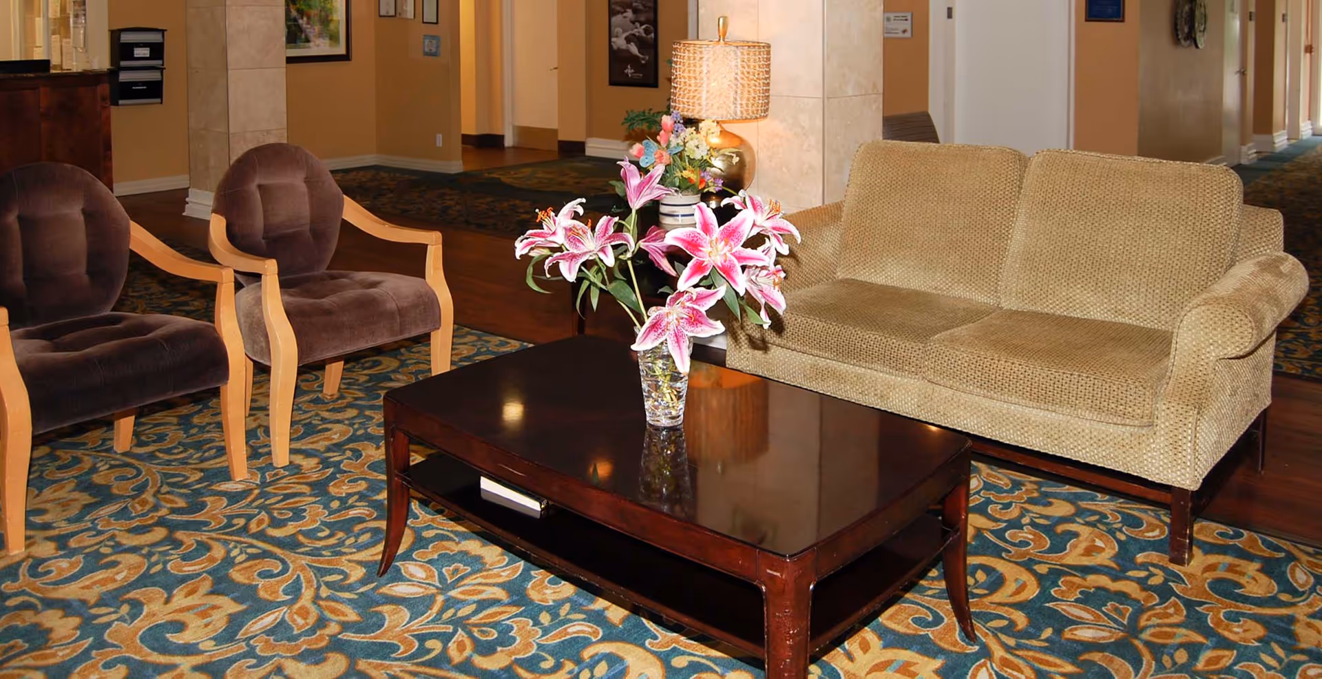 A cozy seating area with a beige couch and two brown cushioned chairs around a dark wooden coffee table with a vase of pink lilies. The area has a patterned carpet and a lamp on a side table in the background.