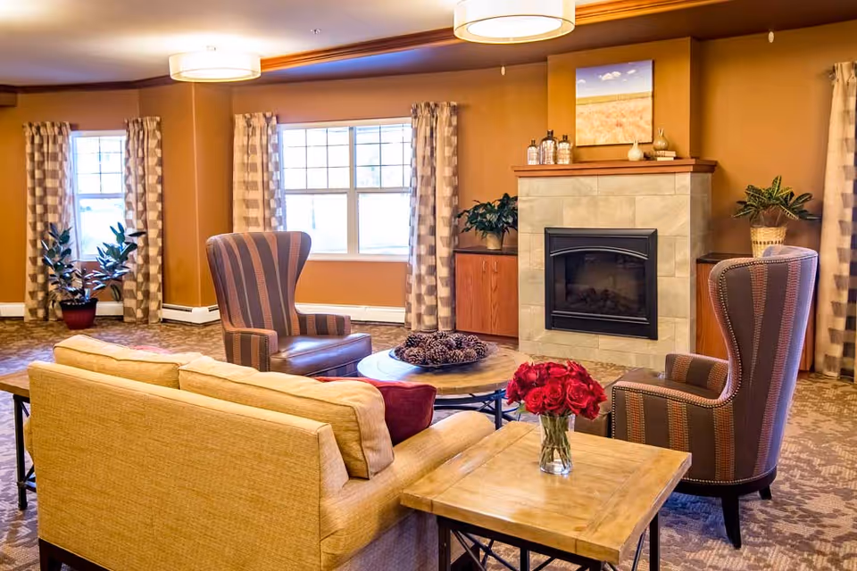 Cozy senior living common room with a sofa, wingback chairs, coffee table, fireplace, and windows.