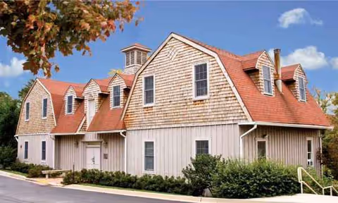 A beige wooden senior living building with red gabled roofs and surrounding greenery.