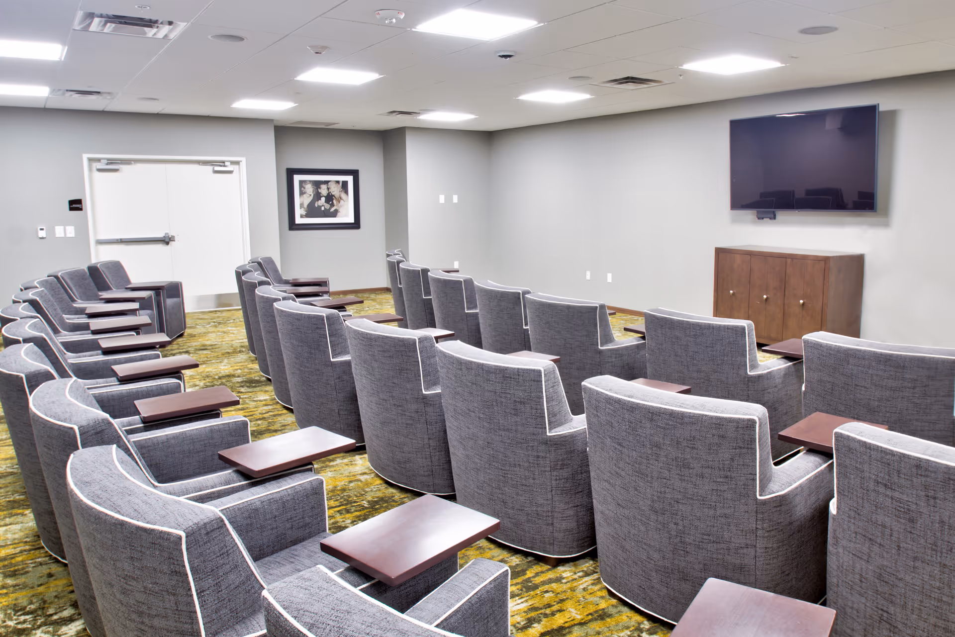 A small theater or media room with multiple rows of gray upholstered chairs, each equipped with a small wooden side table. A flat-screen TV is mounted on the wall in front of the seating area, above a wooden cabinet. The room has gray walls, a patterned green carpet, and a white double door at the back.