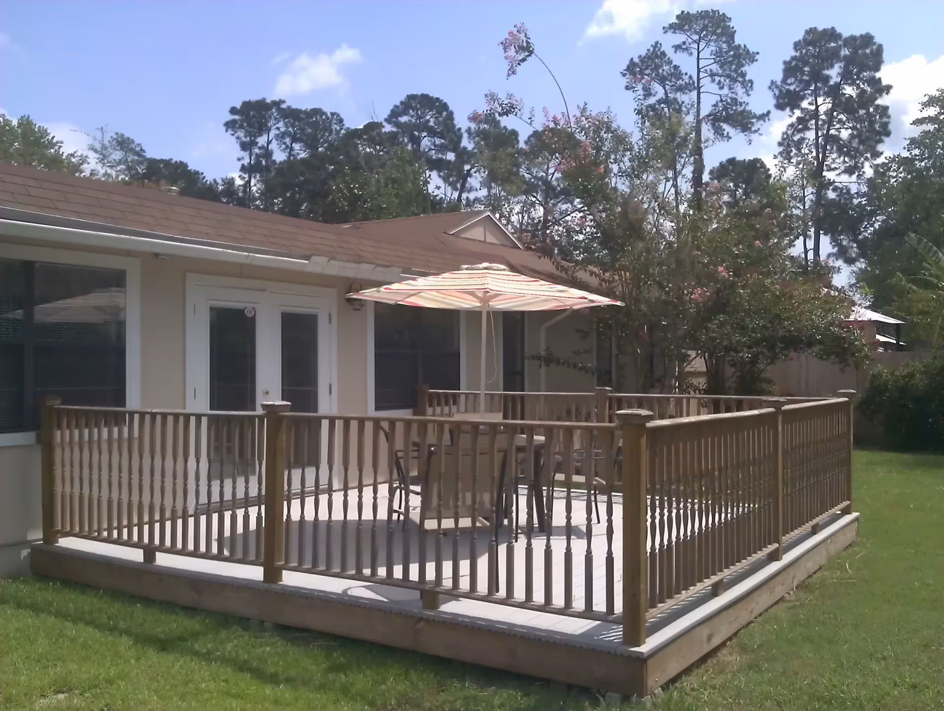 Outdoor patio area with a wooden railing surrounding a raised deck. On the deck, there is a table with several chairs and a large striped umbrella providing shade. The patio is adjacent to a beige building with white-framed double doors and windows. Trees and greenery are visible in the background under a partly cloudy sky.