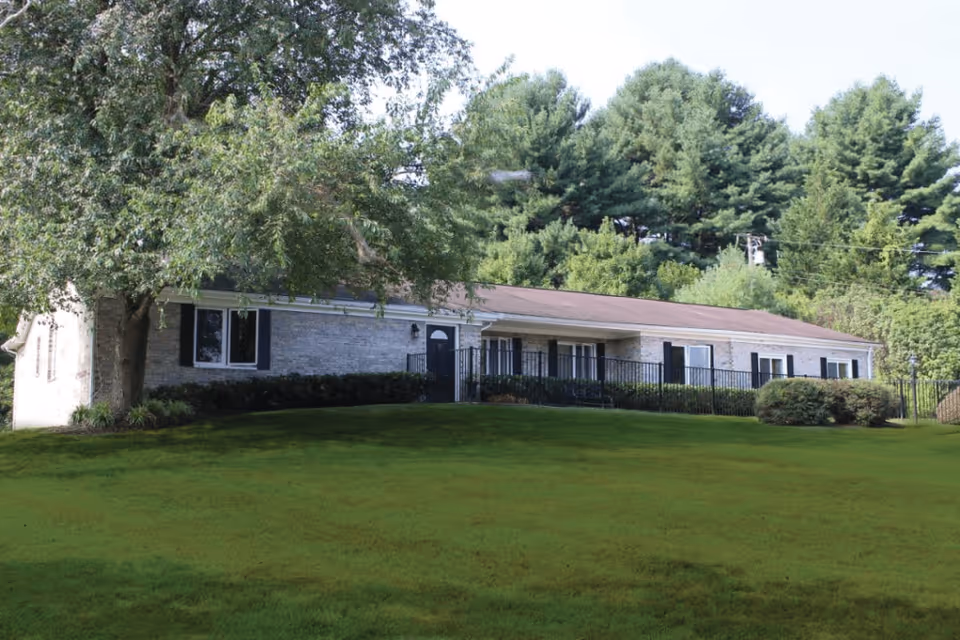 Single-story brick building with multiple windows and a front door, surrounded by green grass and trees under a clear sky.