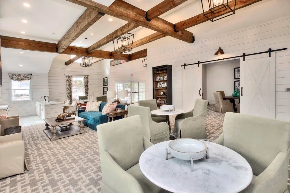 A bright and spacious living room area with exposed wooden ceiling beams, multiple seating arrangements including green armchairs around a round marble table, a teal sofa, beige armchairs, and a wooden coffee table. The room features white shiplap walls, large windows with floral curtains, and sliding barn doors leading to another room with a dining table and chairs.