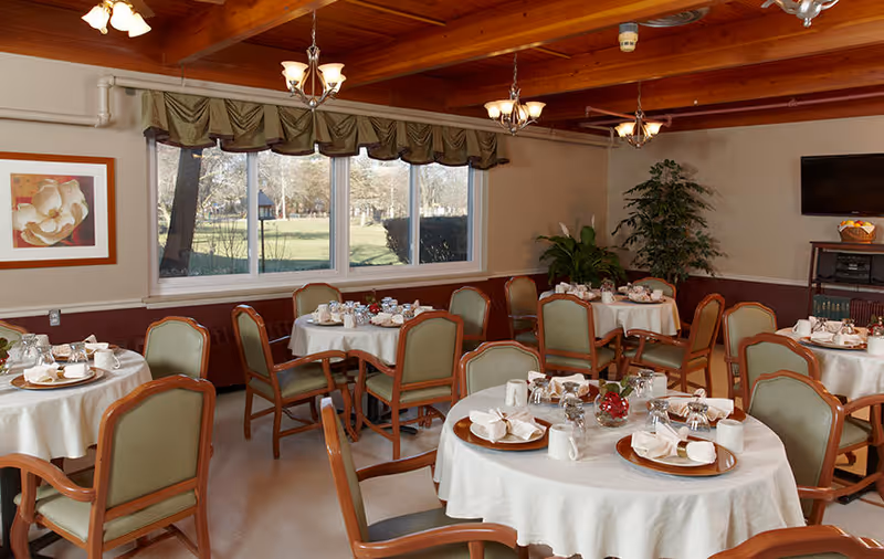 Dining room with round tables covered in white tablecloths, set with plates, cups, napkins, and silverware. The room has green cushioned wooden chairs, large windows with a view of trees and grass outside, wooden ceiling beams, and several light fixtures. There is a framed flower painting on the wall and a TV mounted on the far wall with a small shelf below it holding books and a basket of fruit.