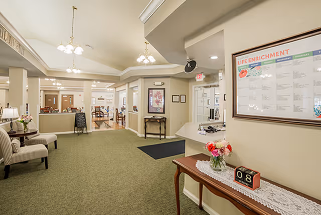 Reception lobby of a senior living facility with seating, a front desk, and a table with flowers beneath a 'Life Enrichment' board.