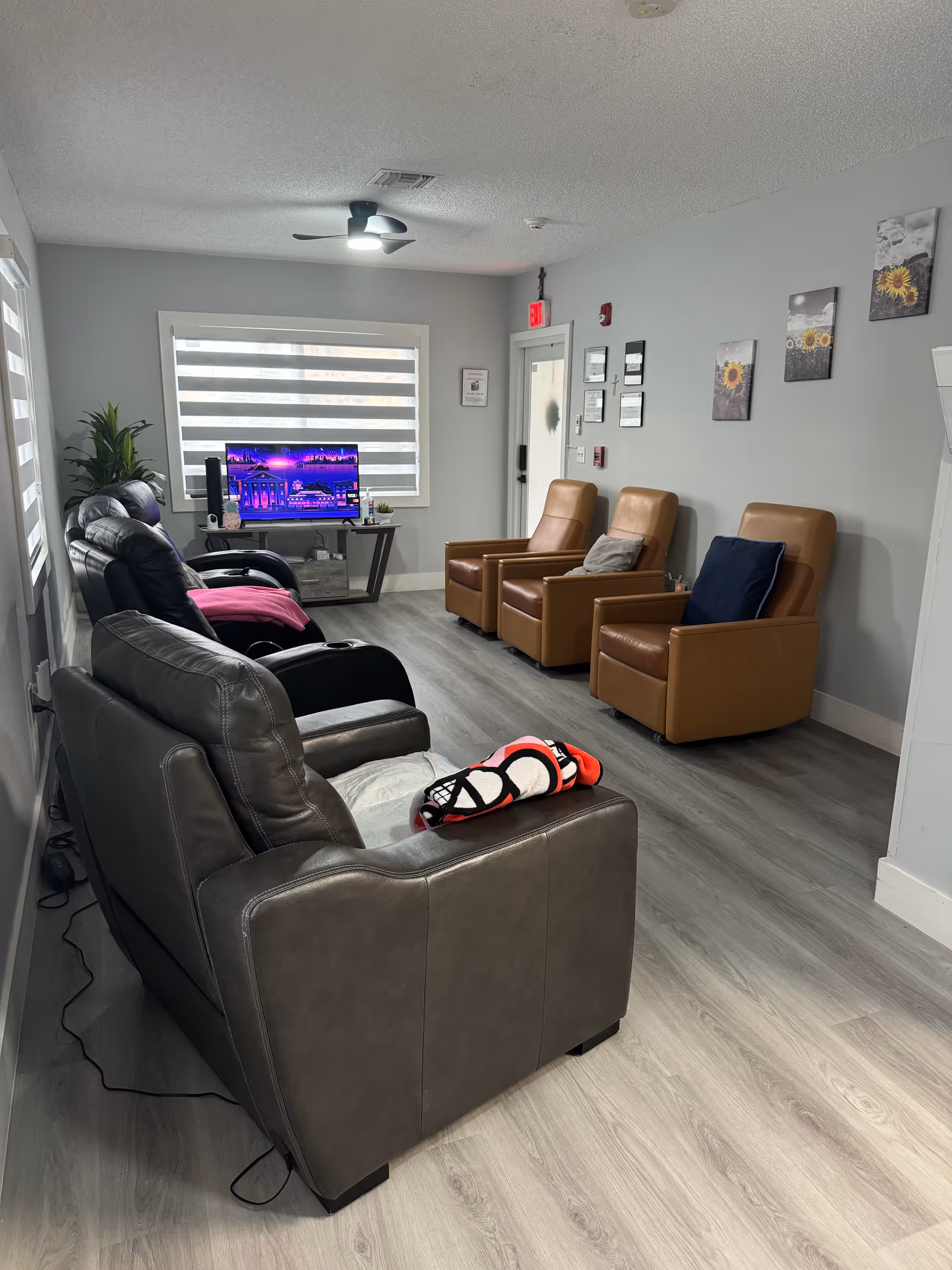 A cozy living room with a row of three brown leather armchairs against a gray wall decorated with sunflower-themed artwork. Opposite the armchairs are two black recliners and a gray recliner, some with blankets on them. A flat-screen TV is on a stand near a window with horizontal blinds, and a green plant is placed beside the TV. The room has light wood flooring and a ceiling fan with a light.