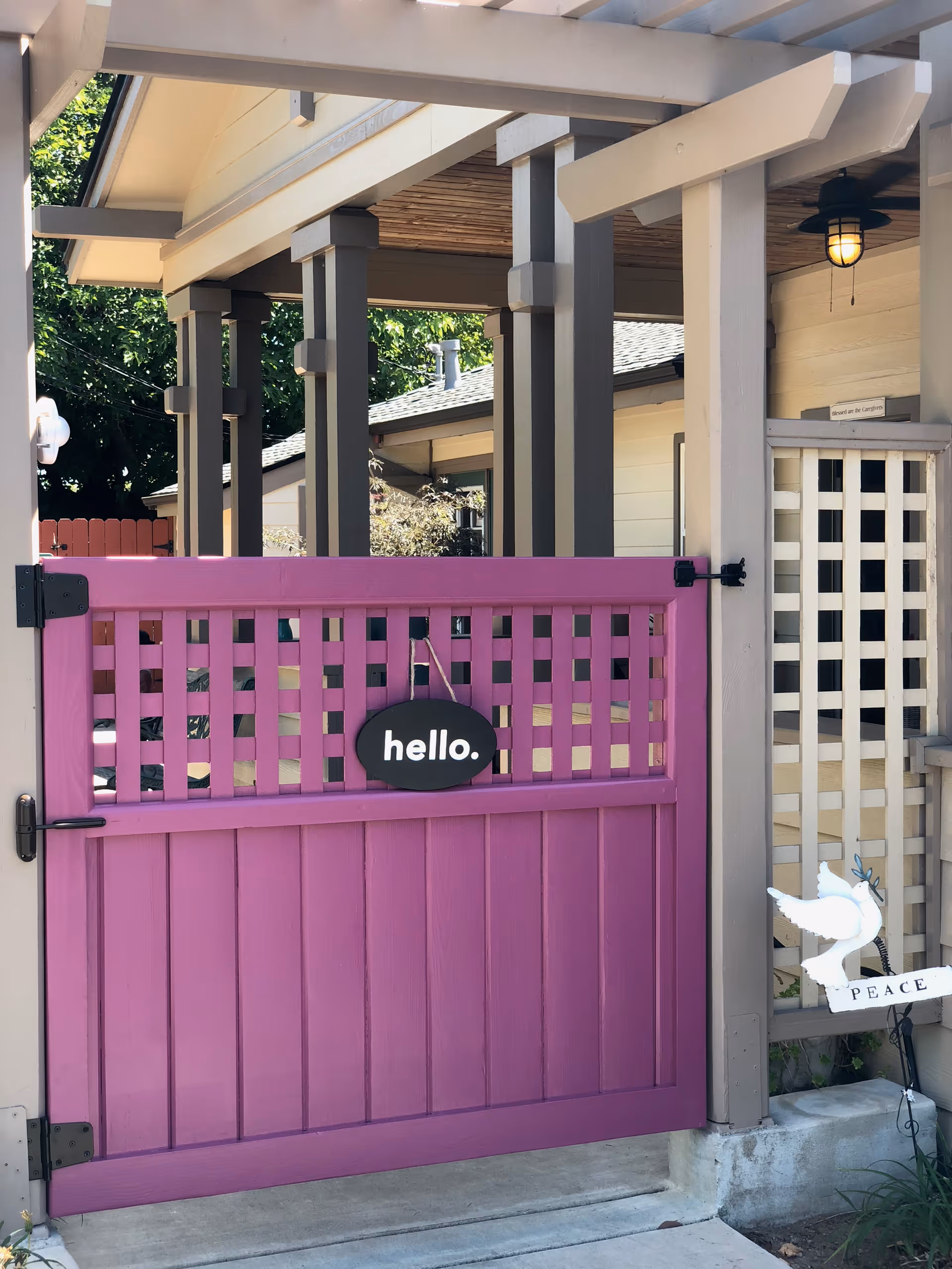 A purple wooden gate with a lattice top and a small black sign hanging on it that says 'hello.' The gate is part of a covered porch area with beige and gray pillars. To the right of the gate, there is a white decorative dove holding an olive branch with a sign that says 'PEACE.' Trees and a house roof are visible in the background.