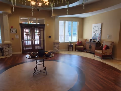 Reception lobby with circular wood-and-tile floor, round table centerpiece, seating chairs by windows, and double glass entrance doors.