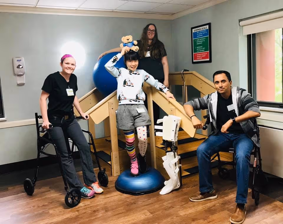 Four people in a rehabilitation room with wooden stairs and exercise equipment. One person is standing on a balance cushion holding a teddy bear, another is sitting on a chair with a leg brace, and two others are standing nearby, one with a walker. The room has light green walls, a window, and a poster on the wall.