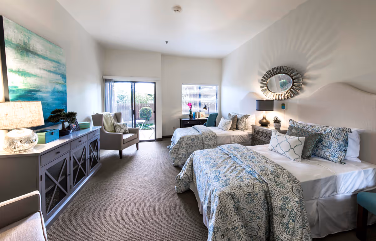 Bright assisted-living bedroom with two twin beds in patterned bedding, a sitting area, console table, and a sliding glass door to a small patio.