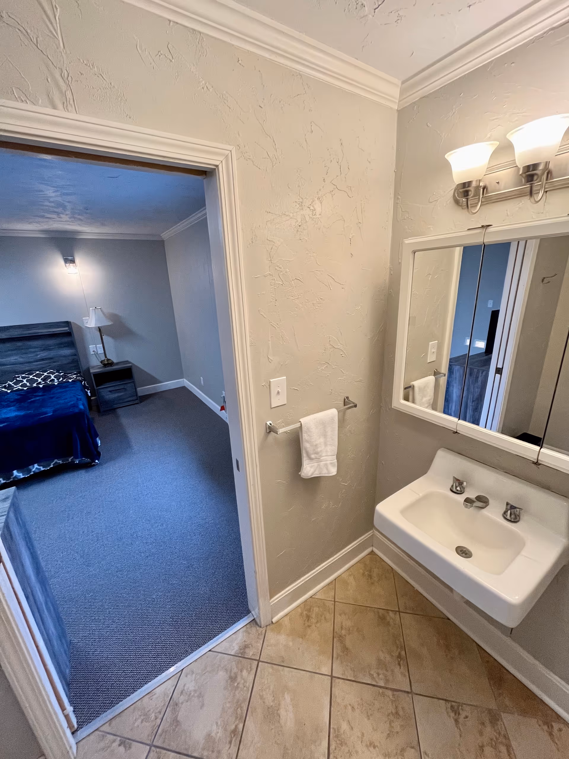 Small bathroom with a wall-mounted sink, mirror, and towel rack, looking into an adjacent bedroom with a bed and nightstand.