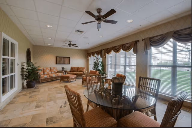 A spacious, well-lit living room area with large windows covered by blinds and valances. The room features a round glass-top table surrounded by wicker chairs with cushions, two ceiling fans, a tiled floor, and a seating area with cushioned wicker sofas and an ottoman. There are also potted plants and wall decor.