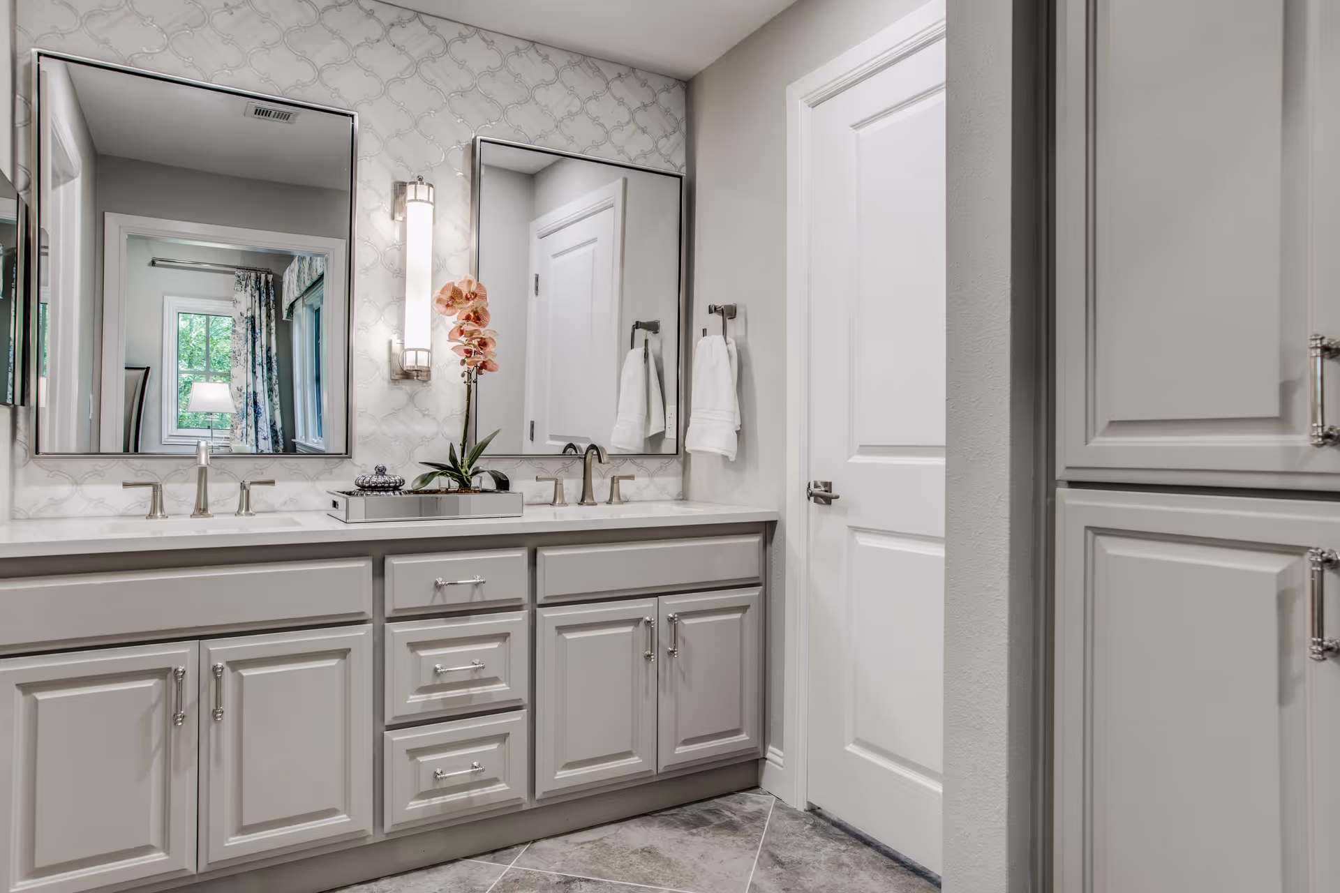 Bright bathroom with a double-sink vanity, two mirrors, gray cabinetry, and decorative orchid on the countertop.