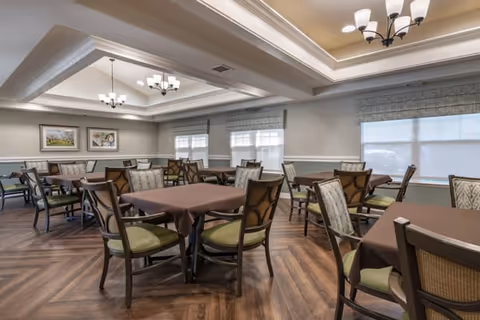 A spacious dining room with multiple tables covered in brown tablecloths and surrounded by cushioned chairs. The room features large windows with blinds, framed artwork on the walls, and elegant ceiling light fixtures. The floor has a wood pattern design.
