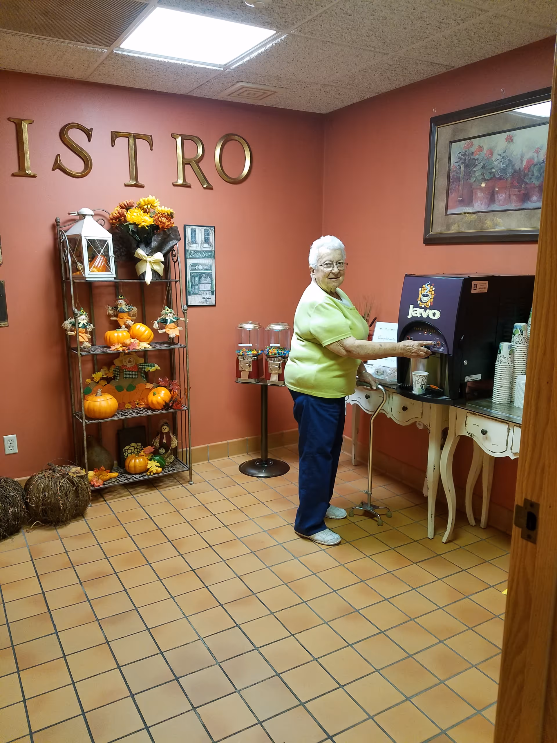 An elderly woman in a green shirt and blue pants is dispensing a drink from a Javo beverage machine in a room with terracotta tiled floor and orange walls. The wall has large gold letters spelling 'BISTRO' and a decorative shelf with autumn-themed decorations including pumpkins and flowers. There is a framed floral painting on the wall above the beverage machine.