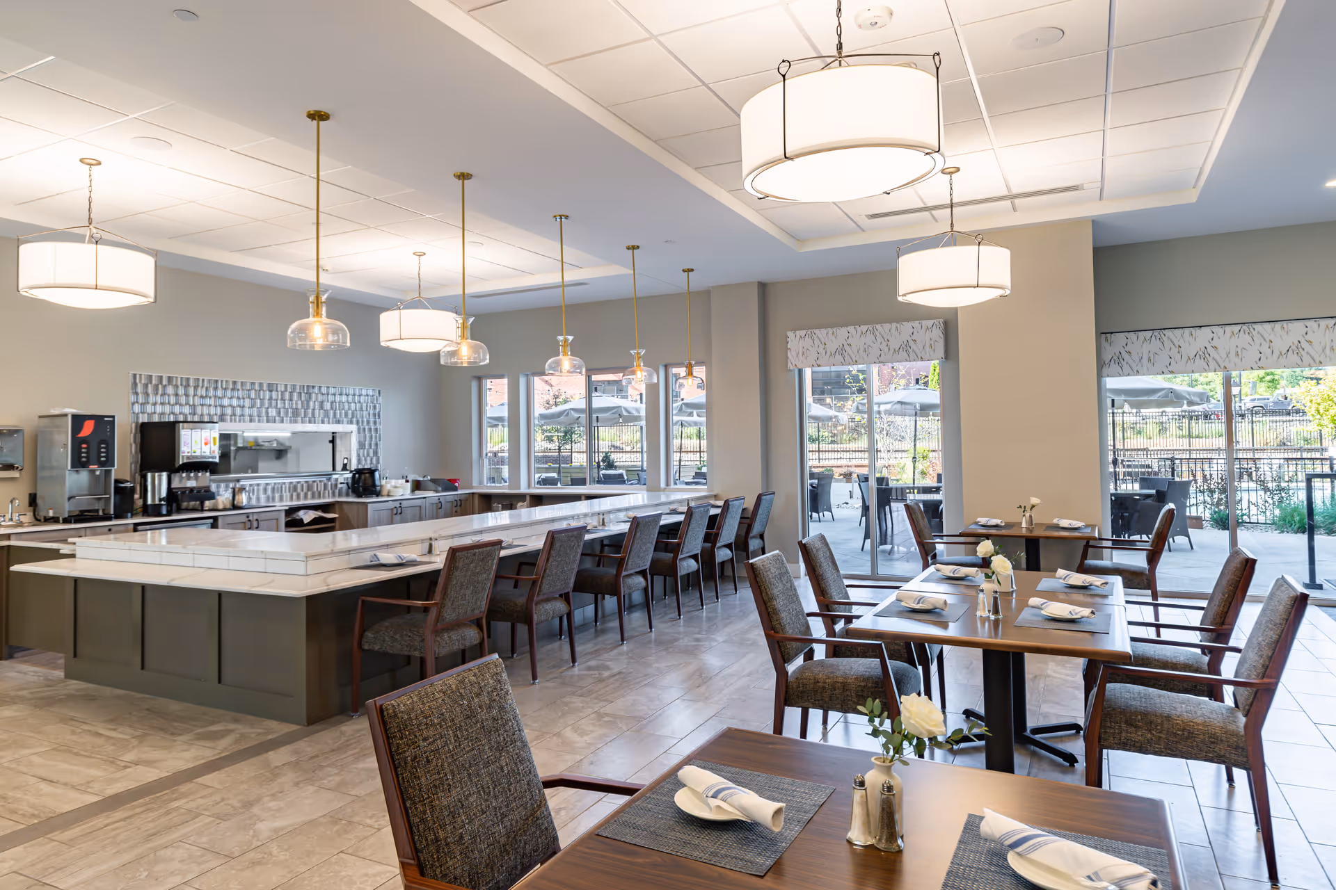 A bright and modern dining room in Merrill Gardens at Greenville featuring a long counter with bar stools, several dining tables set with napkins and silverware, large windows and glass doors leading to an outdoor patio area with tables and umbrellas, and stylish pendant and ceiling lights.