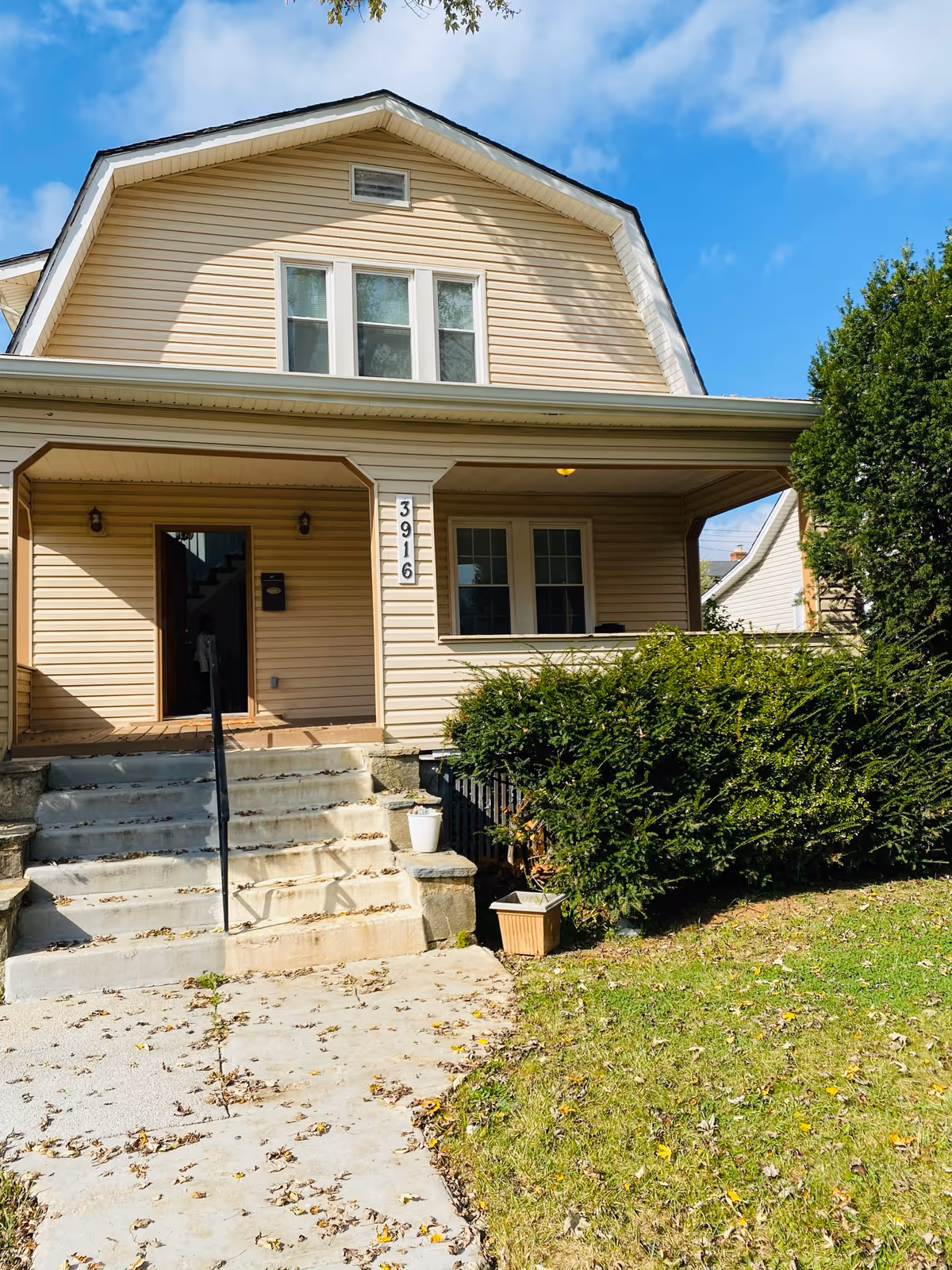 Front exterior view of a beige two-story house with a covered porch, stairs leading up to the front door, and green bushes on the right side under a blue sky with some clouds.