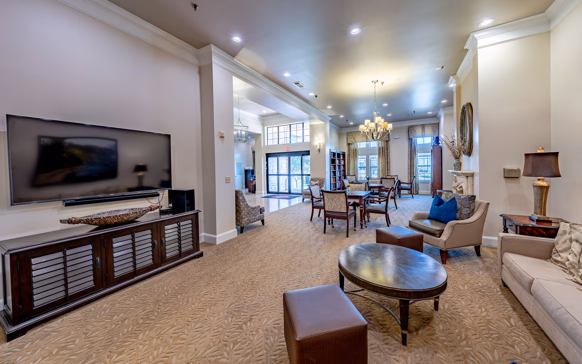A spacious and well-lit living room area in a senior living facility featuring a large flat-screen TV mounted on the wall above a wooden cabinet, comfortable armchairs and sofas with cushions, a round coffee table, and a dining area with tables and chairs near large windows with curtains. The room has beige walls, carpeted floors, recessed lighting, and decorative elements including a chandelier and a wall mirror.