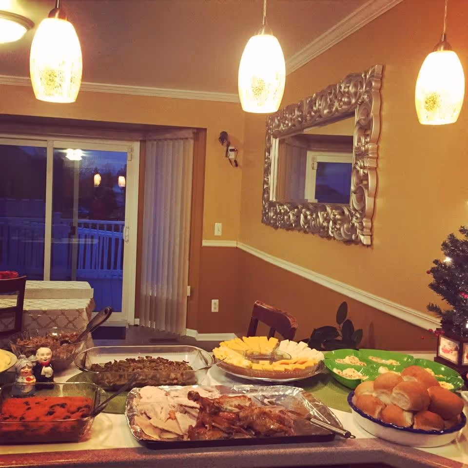 A dining area with a table set with various dishes including sliced turkey, rolls, cheese platter, and other side dishes. The room has warm yellow walls, three hanging pendant lights, a large decorative mirror on the wall, and a sliding glass door with vertical blinds leading to an outdoor balcony or patio.
