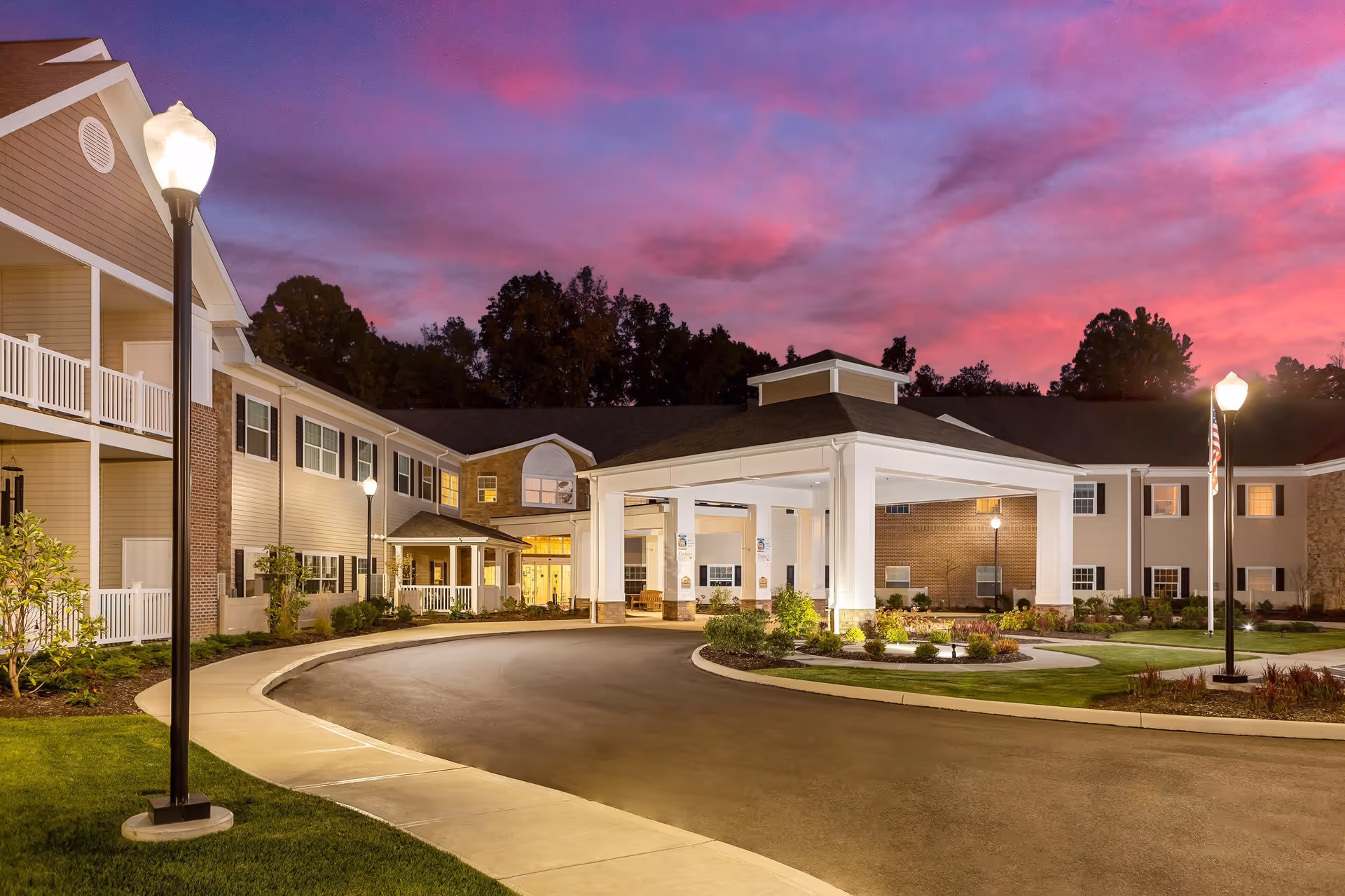 Exterior view of the Danbury Brunswick senior living facility at dusk, showing a large covered entrance with white columns, well-lit street lamps, landscaped greenery, and a vibrant purple and pink sky in the background.