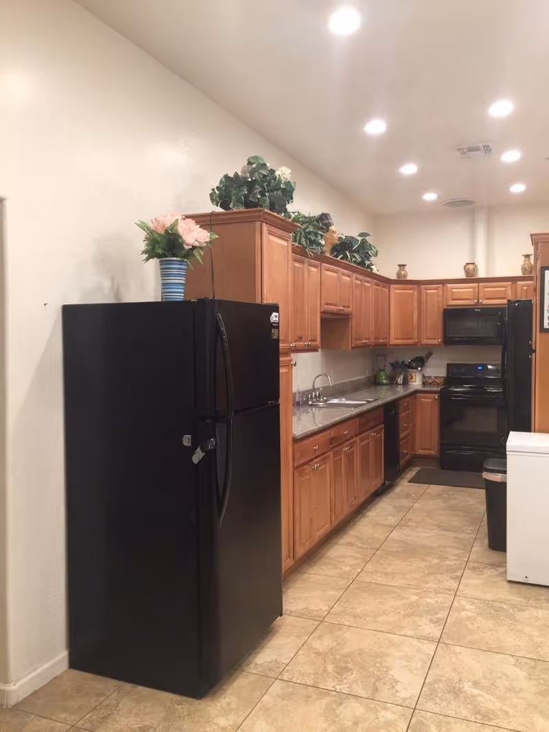 A kitchen with wooden cabinets, a black refrigerator, a black stove with a microwave above it, a double sink, and a countertop with various kitchen items. There are decorative plants on top of the cabinets and tiled flooring.