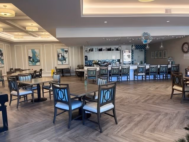 A spacious dining area in an assisted living facility featuring multiple wooden tables and chairs with light blue cushions. The room has a modern design with light wood flooring arranged in a herringbone pattern, white walls with decorative molding, and abstract blue artwork. In the background, there is a kitchen area with a long counter and several high chairs, pendant lights, and kitchen appliances.