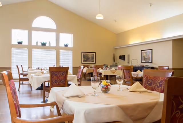 A bright dining room with multiple tables covered in beige tablecloths and set with glasses, napkins, and silverware. The room features large windows with blinds and a high ceiling with a hanging light fixture. There are framed pictures on the walls and wooden chairs with patterned upholstery around the tables.