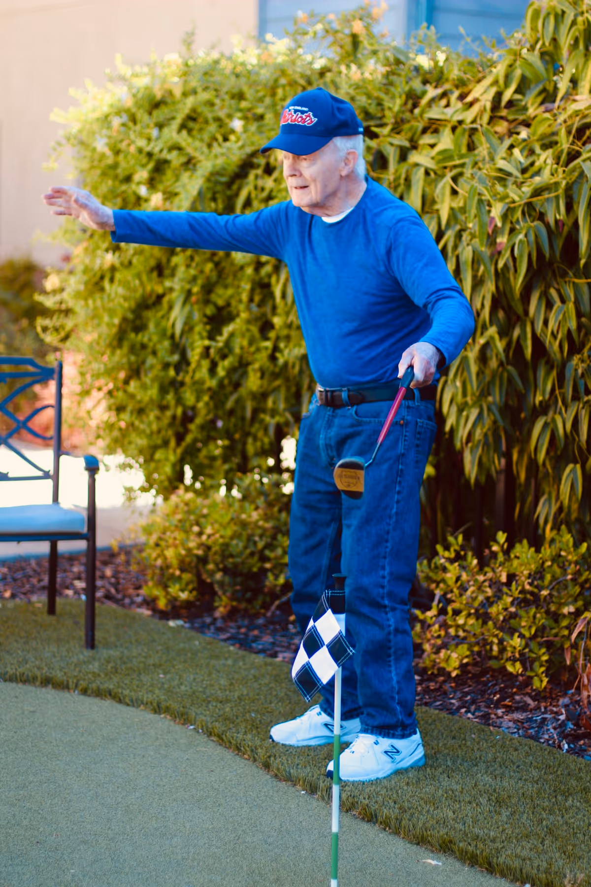 An elderly man wearing a blue long-sleeve shirt, blue jeans, white sneakers, and a blue baseball cap is playing mini golf outdoors. He is holding a putter and appears to be aiming or gesturing towards the hole marked with a checkered flag. There is greenery and a chair in the background.