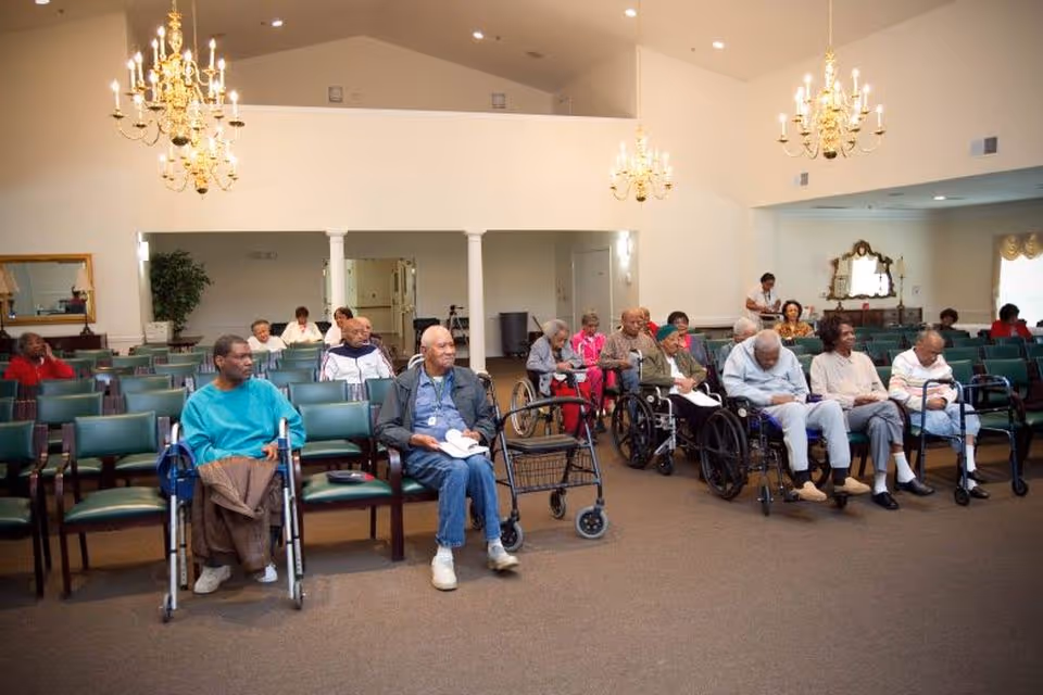 A group of elderly individuals, some in wheelchairs and some seated on chairs, gathered in a spacious room with high ceilings and chandeliers. The room has rows of green chairs and a few people appear to be reading or resting. The setting looks like a common area or meeting room in an assisted living facility.