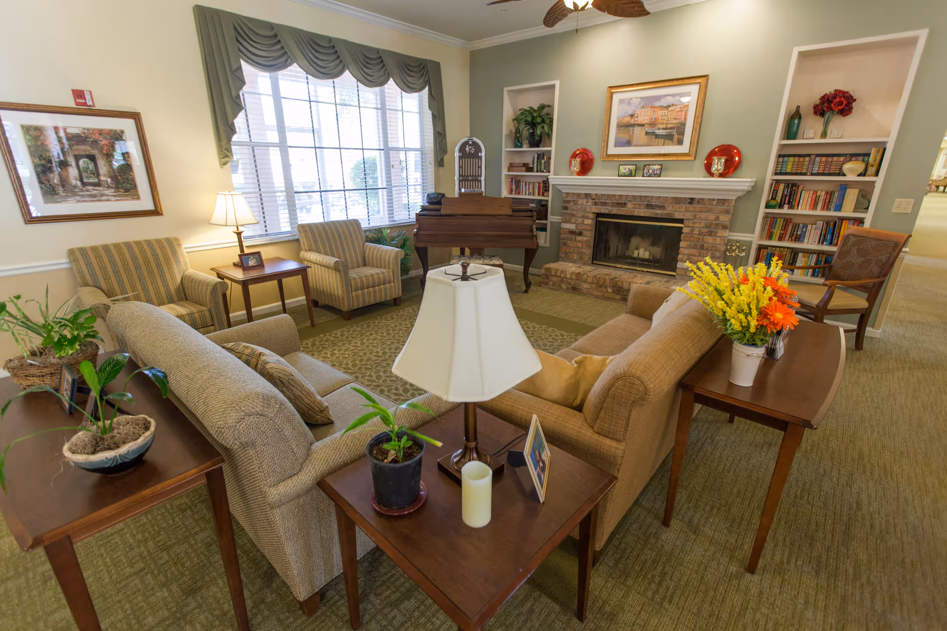 A cozy living room area with two beige sofas, two striped armchairs, a wooden coffee table with a lamp and plants, a brick fireplace with decorative items on the mantel, built-in bookshelves filled with books and decor, a large window with green valance curtains, a framed painting on the wall, and a piano in the corner.