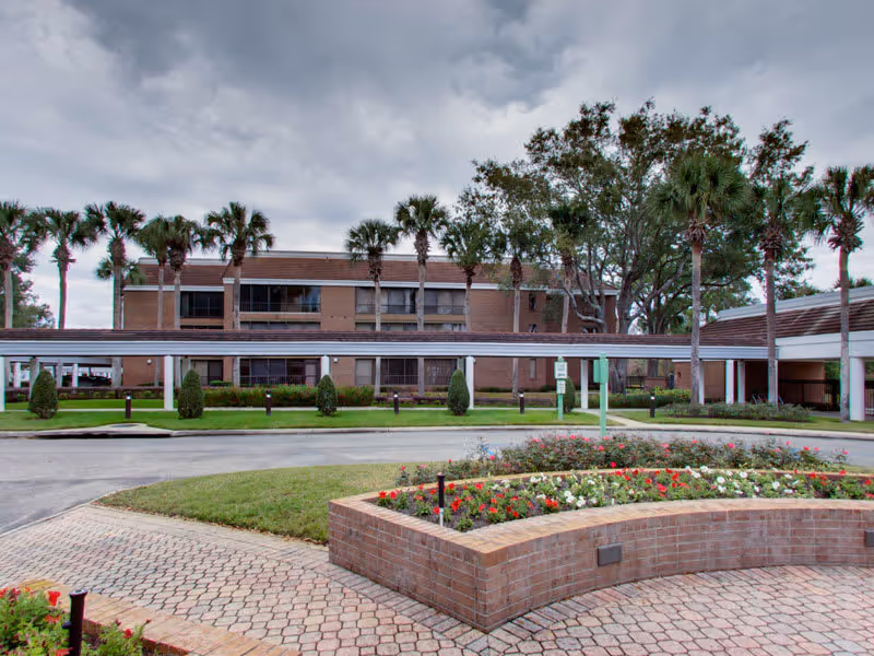 Exterior view of a senior living facility named Village on the Green, showing a brick building with multiple windows, palm trees, and a covered walkway. In the foreground, there is a circular brick planter with colorful flowers and a paved driveway under a cloudy sky.