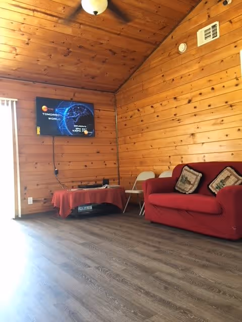 Interior of a room with wooden paneled walls and ceiling, a mounted flat-screen TV displaying a digital globe, a red couch with two decorative pillows, a small table covered with a red cloth, and a white folding chair.