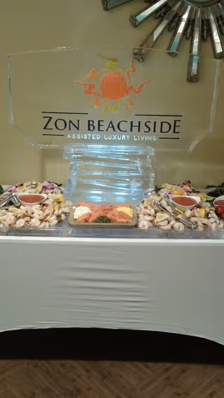 Ice sculpture sign reading "ZON BEACHSIDE ASSISTED LUXURY LIVING" displayed above a seafood buffet with shrimp and dipping sauces.
